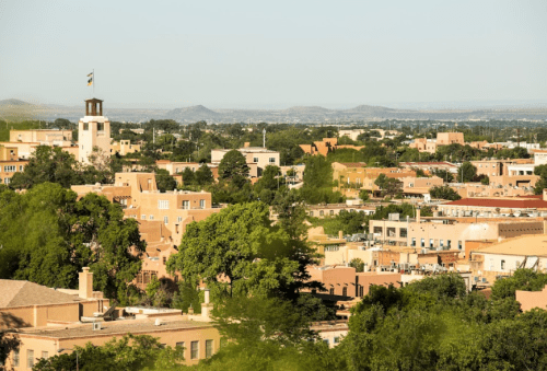 A panoramic view of a city with adobe-style buildings, greenery, and distant mountains under a clear blue sky.