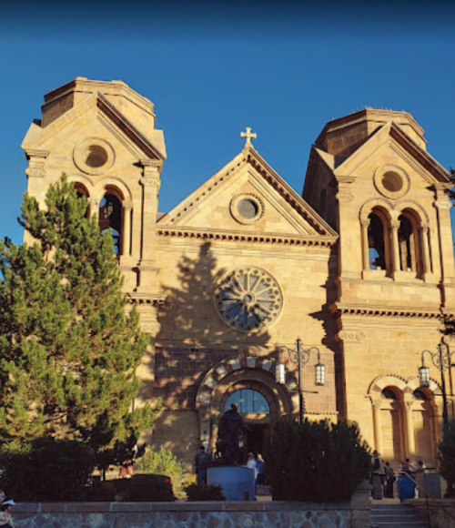 Historic stone church with twin towers and a large rose window, surrounded by trees and people in the foreground.