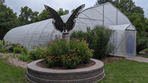 A greenhouse with a butterfly sculpture in front, surrounded by colorful flowers and greenery.