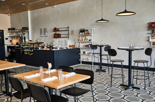 Modern restaurant interior with wooden tables, black chairs, and a stylish bar area against a gray wall.