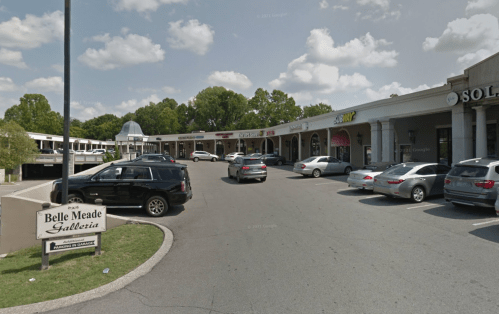 A shopping plaza with several stores, cars parked, and trees in the background under a partly cloudy sky.