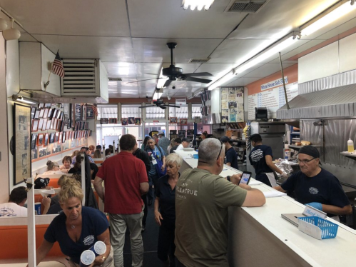 A busy diner with patrons and staff, featuring a counter, kitchen area, and walls adorned with photos and memorabilia.