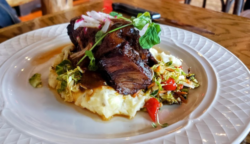 A plate of tender beef on creamy mashed potatoes, topped with radish and microgreens, accompanied by sautéed vegetables.