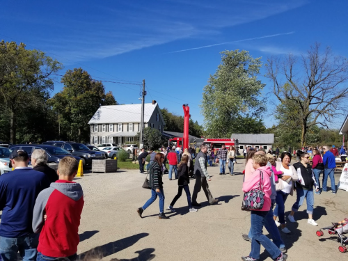 A busy outdoor market scene with people walking, cars parked, and a large red structure in the background under a clear blue sky.