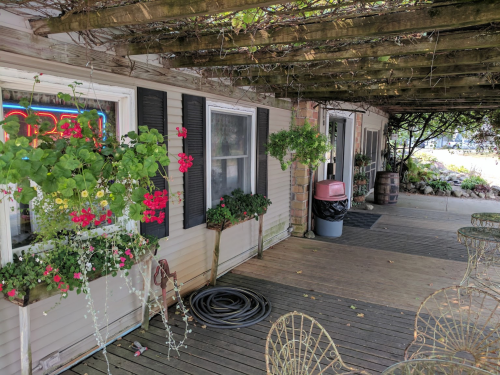 A cozy outdoor area with a wooden deck, potted flowers, and a shop entrance under a vine-covered pergola.