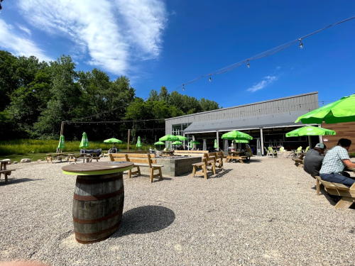 Outdoor seating area with wooden tables and green umbrellas, surrounded by trees and a blue sky.