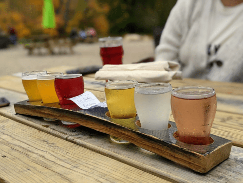 A wooden flight board with eight small glasses of various colorful beers on a rustic table outdoors.