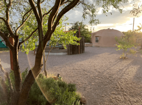 A serene outdoor space featuring a tree, gravel ground, and a round adobe structure under a cloudy sky.