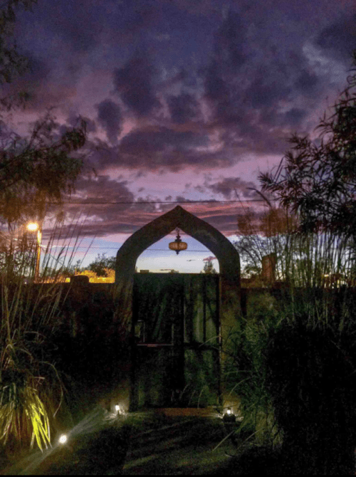 A dark archway leads to a colorful sunset sky, framed by tall grasses and soft lighting along the path.