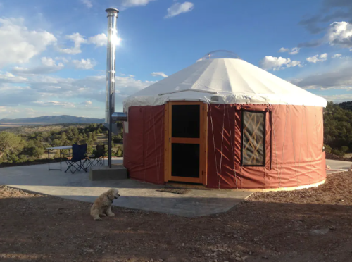 A yurt with a chimney sits on a rocky landscape, with two chairs nearby and a dog resting in front.