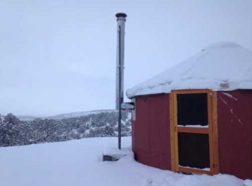 A red yurt covered in snow, with a chimney, set against a snowy landscape and overcast sky.