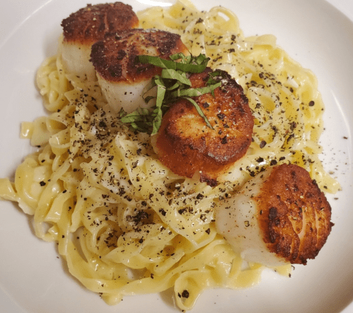 A plate of fettuccine topped with seared scallops and fresh basil, sprinkled with black pepper.