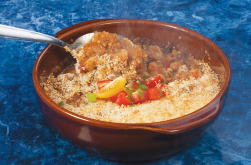 A brown bowl filled with a savory dish topped with breadcrumbs, diced tomatoes, and green peppers, with steam rising.