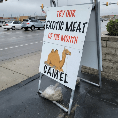Sign advertising "Exotic Meat of the Month: Camel" with a camel illustration, placed outside in a parking lot.