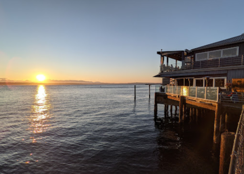 Sunset over calm waters beside a wooden pier and a restaurant, with mountains in the distance.