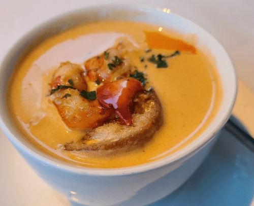 A bowl of creamy lobster bisque with chunks of lobster and a drizzle of oil, garnished with herbs.