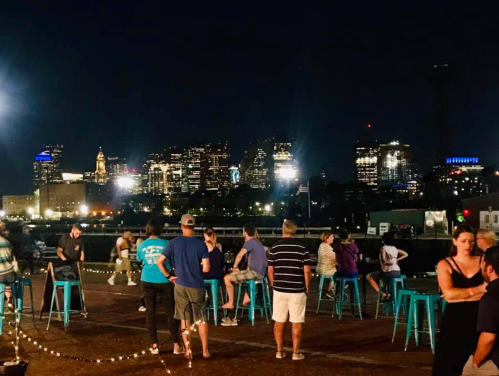 A lively rooftop scene at night with people socializing and a city skyline illuminated in the background.