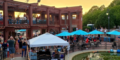 A lively outdoor event at sunset, with people gathered around a stage and blue umbrellas by a pond.