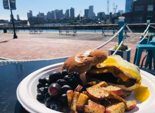 A breakfast plate with a burger topped with an egg, alongside berries and roasted potatoes, with a city skyline in the background.