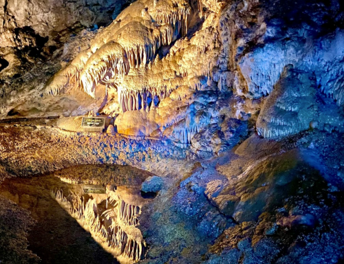 Stalactites and stalagmites illuminated in a cave, reflecting in a still pool of water.