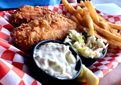A plate of crispy fried fish fillets, French fries, coleslaw, and tartar sauce on a red and white checkered tray.