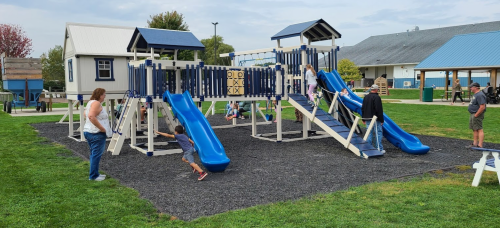 Children play on a blue and white playground with slides and climbing structures, surrounded by grass and adults nearby.