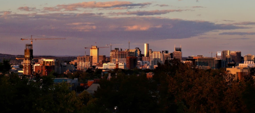 City skyline at sunset with construction cranes and buildings, surrounded by trees and a colorful sky.