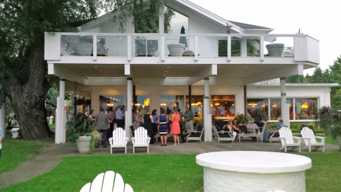 A gathering of people outside a modern building with large windows, surrounded by greenery and white chairs.