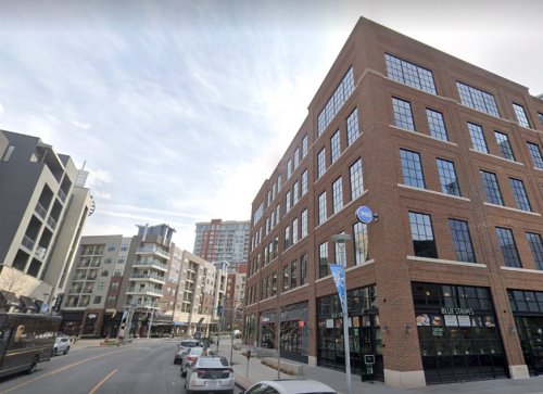 A street view of a brick building with large windows, surrounded by modern buildings and parked cars.