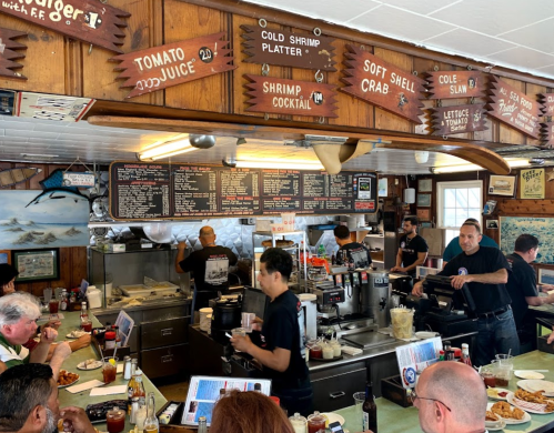 A busy seafood restaurant with patrons at tables and staff serving food, featuring a menu on the wall and a lively atmosphere.