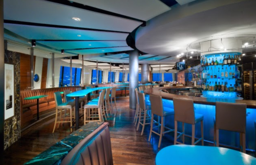 A modern bar with blue lighting, sleek furniture, and a circular design, featuring a well-stocked backlit bar.