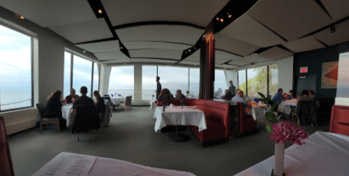 A restaurant interior with diners enjoying views of the ocean through large windows. Tables are set with white linens.