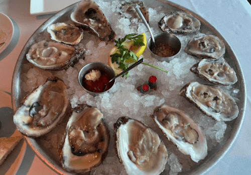 A platter of fresh oysters on ice, garnished with lemon, sauces, and herbs.