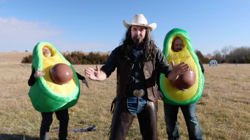 Three people in inflatable avocado costumes stand in a field, with one man in a cowboy hat and leather vest in front.