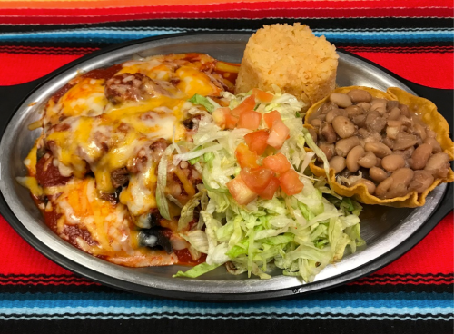 A metal plate with enchiladas topped with cheese, rice, beans, lettuce, and tomatoes, set on a colorful woven tablecloth.