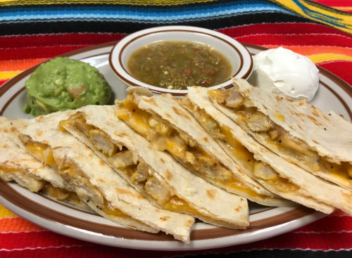 A plate of quesadillas filled with cheese and chicken, served with guacamole, salsa, and sour cream on a colorful tablecloth.