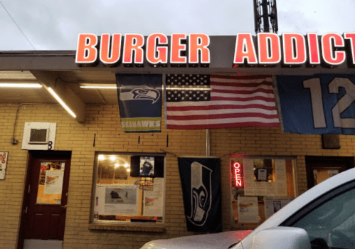 Exterior of "Burger Addict" restaurant with flags, including the American flag and Seattle Seahawks banners, and an "Open" sign.