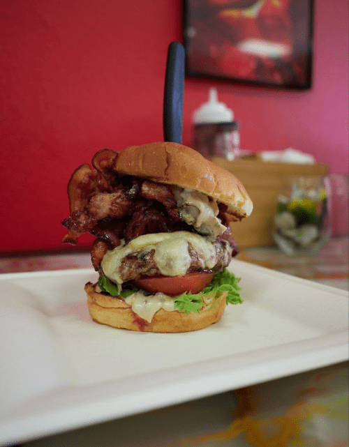A stacked burger with bacon, melted cheese, lettuce, and tomato on a plate, set against a red wall.