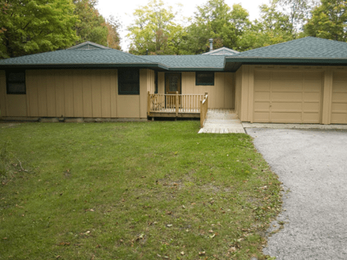 A single-story house with a green roof, wooden deck, and garage, surrounded by grass and trees.