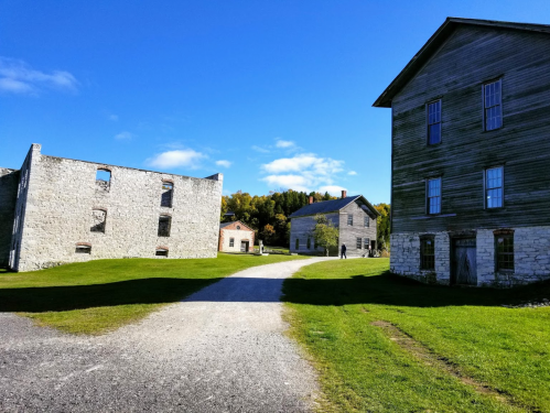 Historic stone and wooden buildings along a gravel path, surrounded by green grass and a clear blue sky.