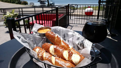 A basket of pretzel sticks with cheese dip and a dark beverage, set against a scenic waterfront view.
