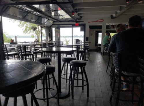 Interior of a restaurant with black tables and stools, large windows showing a lake view, and a few patrons seated.