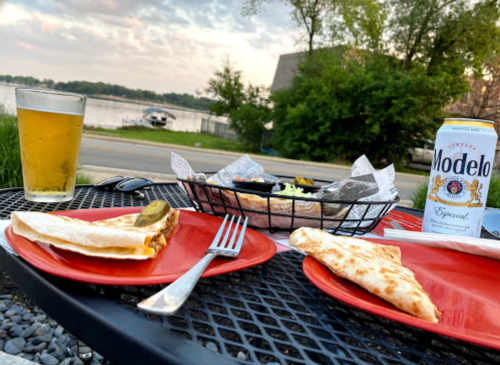 A table by the water with two plates of food, a basket of salad, a beer, and a can of Modelo.
