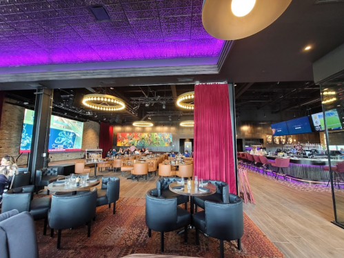 Modern restaurant interior with purple lighting, circular chandeliers, and a bar area, featuring tables and a vibrant mural.