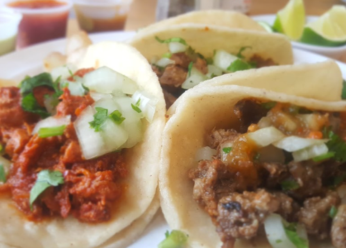 Three tacos on a plate, filled with meat, onions, and cilantro, accompanied by lime wedges and sauces in the background.