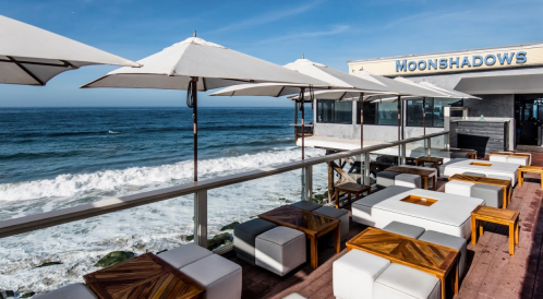 Outdoor seating area with white furniture and umbrellas overlooking the ocean at Moonshadows restaurant.
