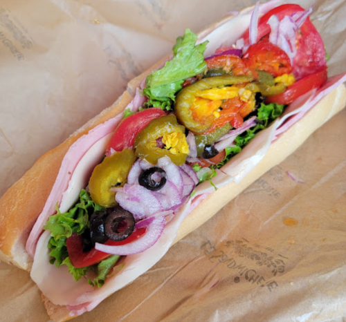 A sub sandwich filled with ham, lettuce, tomatoes, jalapeños, olives, and onions on a toasted hoagie roll.