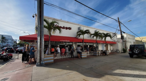 A lively restaurant with outdoor seating, palm trees, and patrons enjoying the sunny day.