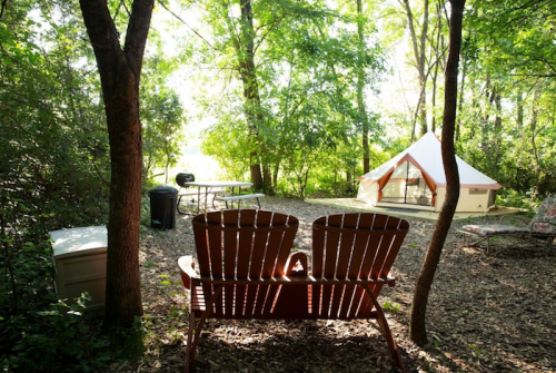 A serene campsite with two wooden chairs, a tent, picnic table, and lush greenery in the background.