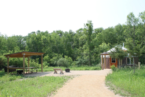 A cozy yurt beside a picnic area, surrounded by lush green trees and a gravel path.
